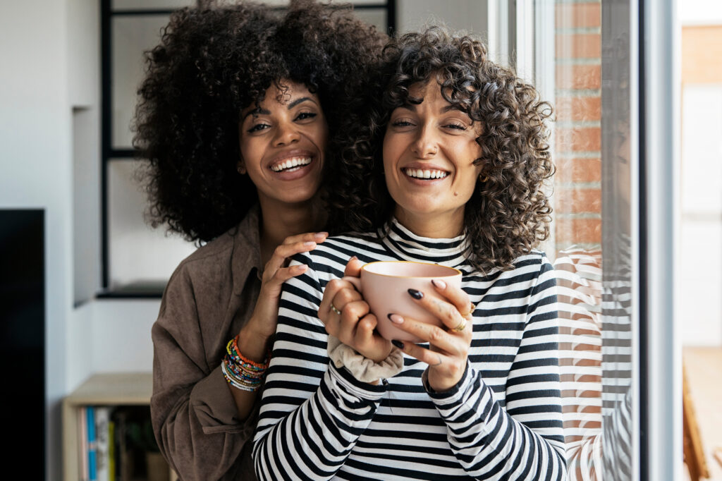 Stock photo of two women having a cup of coffee at home. They are smiling and sharing happy moments. The women look positive and happy. Friendship concept.